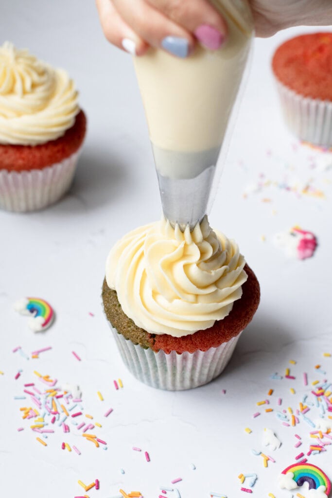 Piping white chocolate buttercream onto rainbow cupcakes