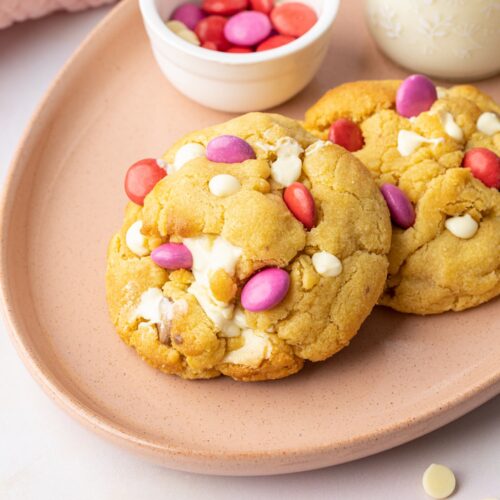 Two thick NYC-style chocolate chunk cookies on a pink plate with a bowl of pink and red Smarties nearby