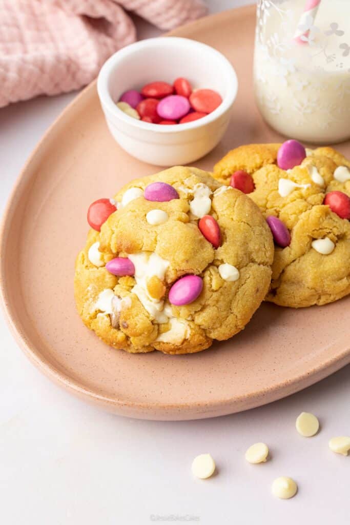Two thick NYC-style chocolate chunk cookies on a pink plate with a bowl of pink and red Smarties nearby