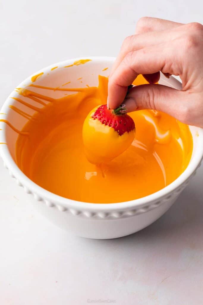 A hand dipping a strawberry into some orange melted chocolate