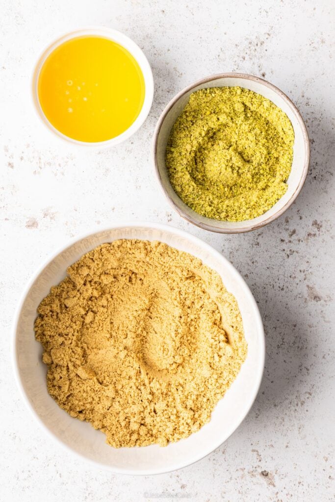 A top down view of 3 bowls, ingredients for pistachio biscuit cheesecake base